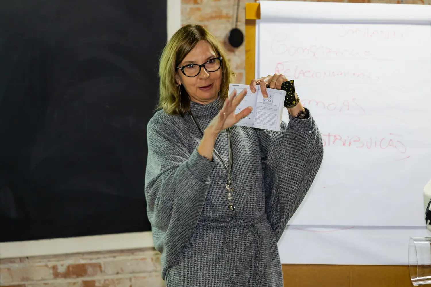 Mentora mostrando um material de prática em aula, ao lado do flip chart escrito à mão com consciência, relacionamento, vendas e distribuição.